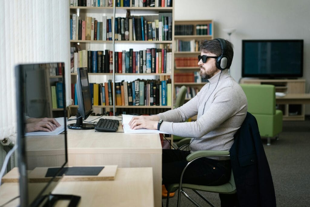 pexels-photo-6981085-6981085 A blind man uses a computer with headphones in a library, highlighting technology's role in accessibility.