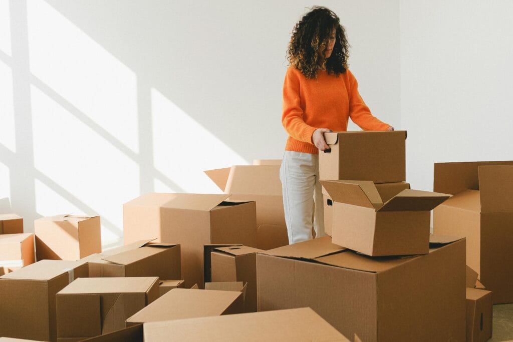 pexels-photo-7203814-7203814 Focused young female with curly dark hair in casual outfit arranging pile of cardboard boxes while preparing for relocation in new apartment