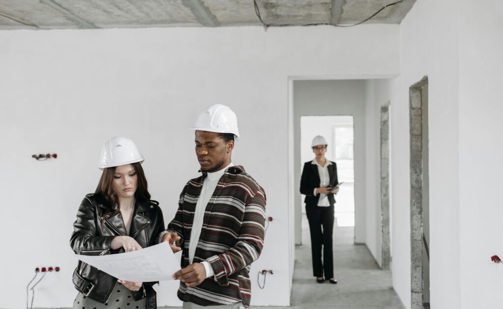 A couple wearing hard hats examines building plans inside an unfinished property with a realtor observing.