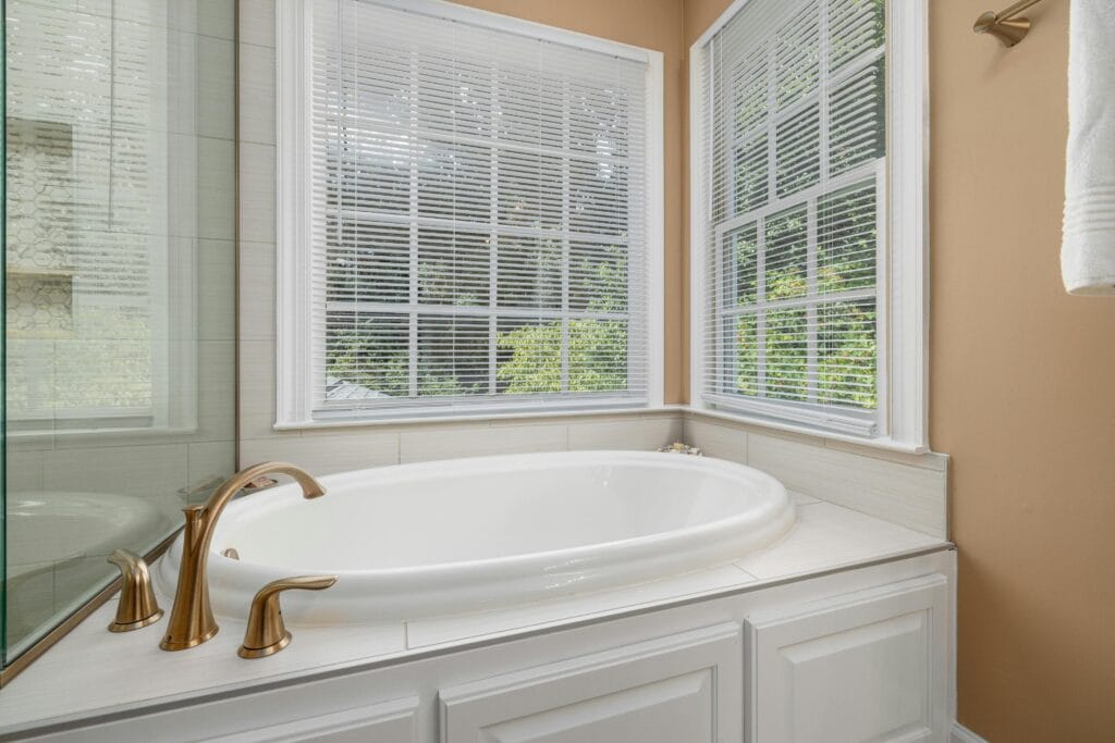 pexels-photo-12908650-12908650 Bright bathroom featuring a white bathtub, brass faucets, and large windows with blinds.