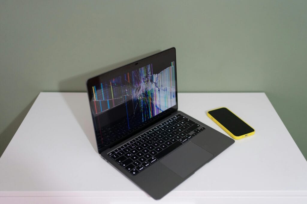 pexels-photo-28379994-28379994 A damaged laptop screen with colorful lines and a yellow smartphone on a white desk.