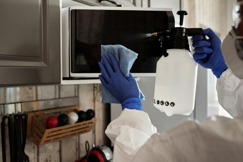 pexels-photo-4099082-4099082 Individual in protective gear cleaning microwave using spray bottle and cloth indoors.