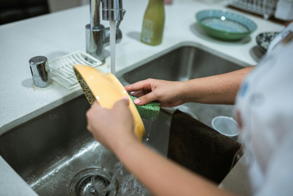 pexels-photo-5591636-5591636 Close-up of hands washing dishes in a kitchen sink with running water.