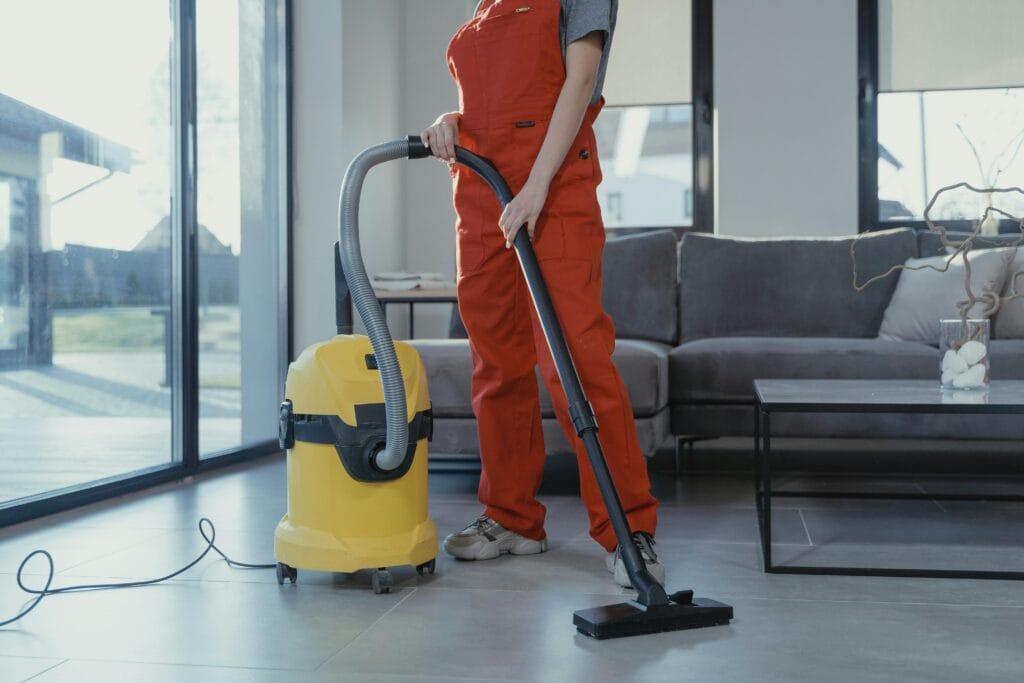 pexels-photo-6195275-6195275 A professional cleaner in red coveralls vacuuming a modern living room with a yellow vacuum cleaner.