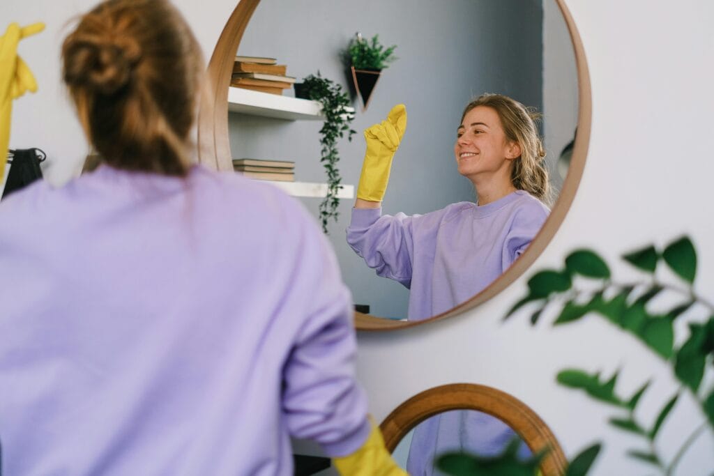 pexels-photo-7513029-7513029 Smiling woman cleaning mirror with yellow gloves indoors, reflecting her image.