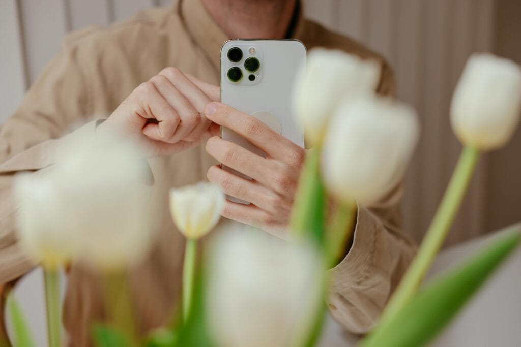 pexels-photo-7657752-7657752 Close-up of a man photographing white tulips indoors with a smartphone.